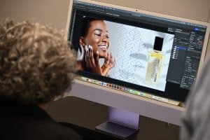 A person with curly hair views a computer screen displaying an image editing program, showing a smiling woman applying cream and a skincare product bottle on the right side.